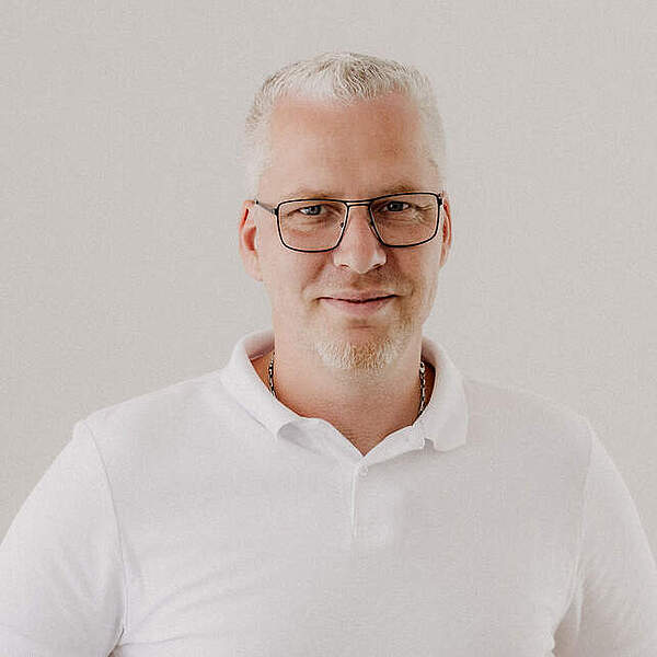 Kurzhaariger Mann mit weißem Dreitagebart und rechteckiger Brille trägt weißes Polohemd vor hellem Hintergrund