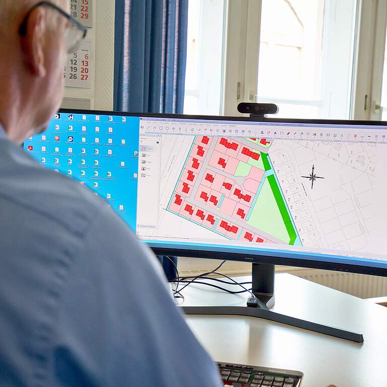 Person sitzt am Schreibtisch vor breitem Monitor und betrachtet farbige Bebauungspläne mit roten Häusern und grünen Flächen