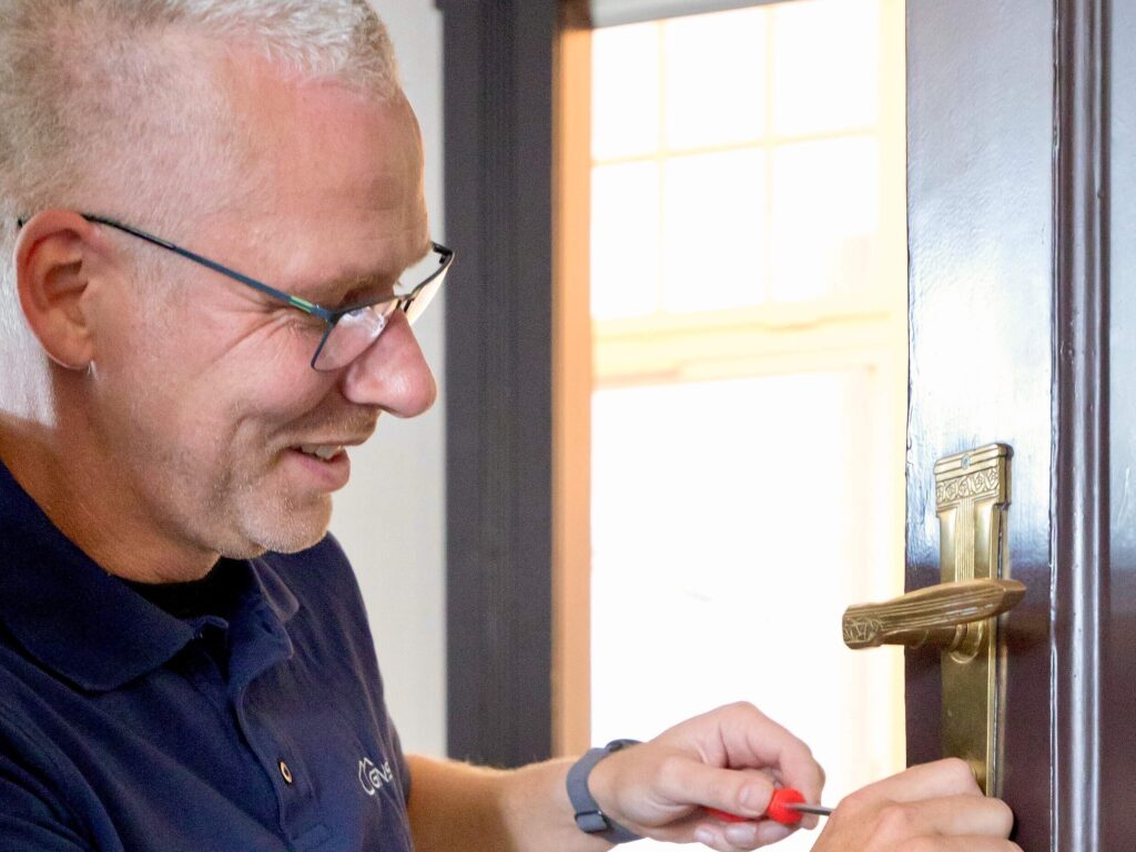 Ein Handwerker mit Brille und dunkelblauem Polo repariert mit einem Schraubendreher einen verzierten Messing-Türgriff an einer Holztür