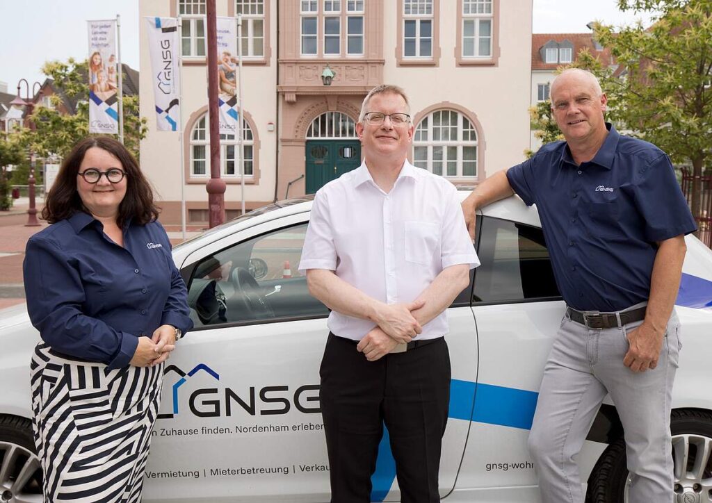 Drei lächelnde GNSG-Mitarbeitende stehen neben einem weißen Firmenwagen vor einem historischen Rathausgebäude