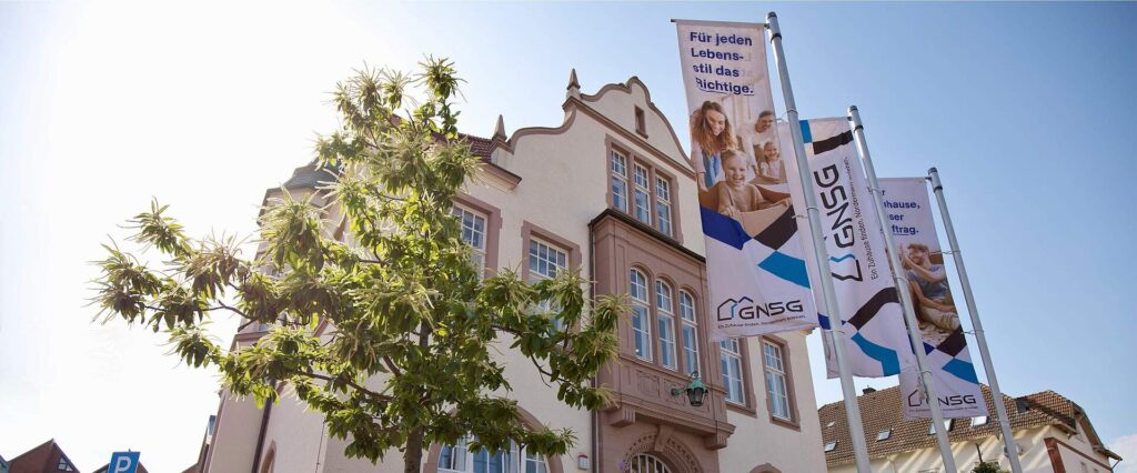 Historisches Verwaltungsgebäude hinter einem jungen Baum, flankiert von Fahnen mit GNSG-Logo und Familienmotiven
