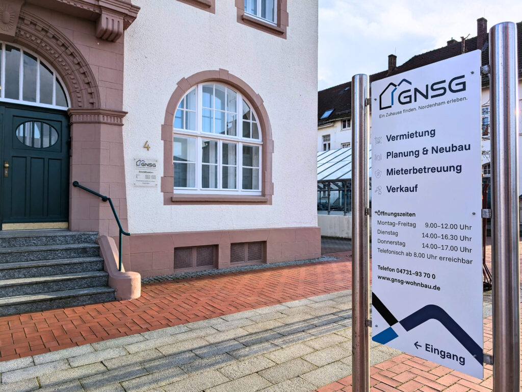 GNSG-Hinweisschild vor Haus mit Angeboten zu Vermietung, Neubau, Mieterbetreuung, Verkauf, Öffnungszeiten und Eingangspfeil